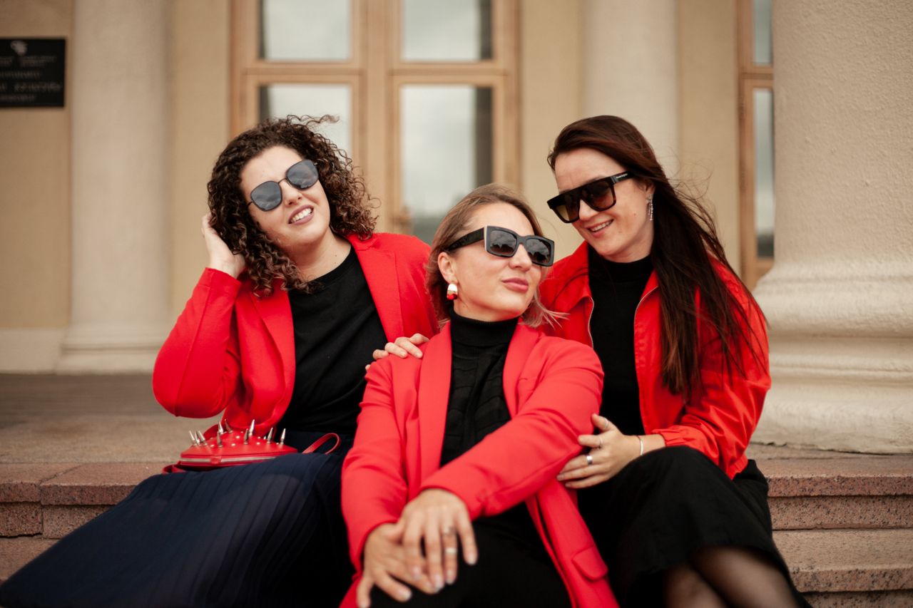 team-01 Three stylish women in red jackets sitting outdoors, enjoying the day together.